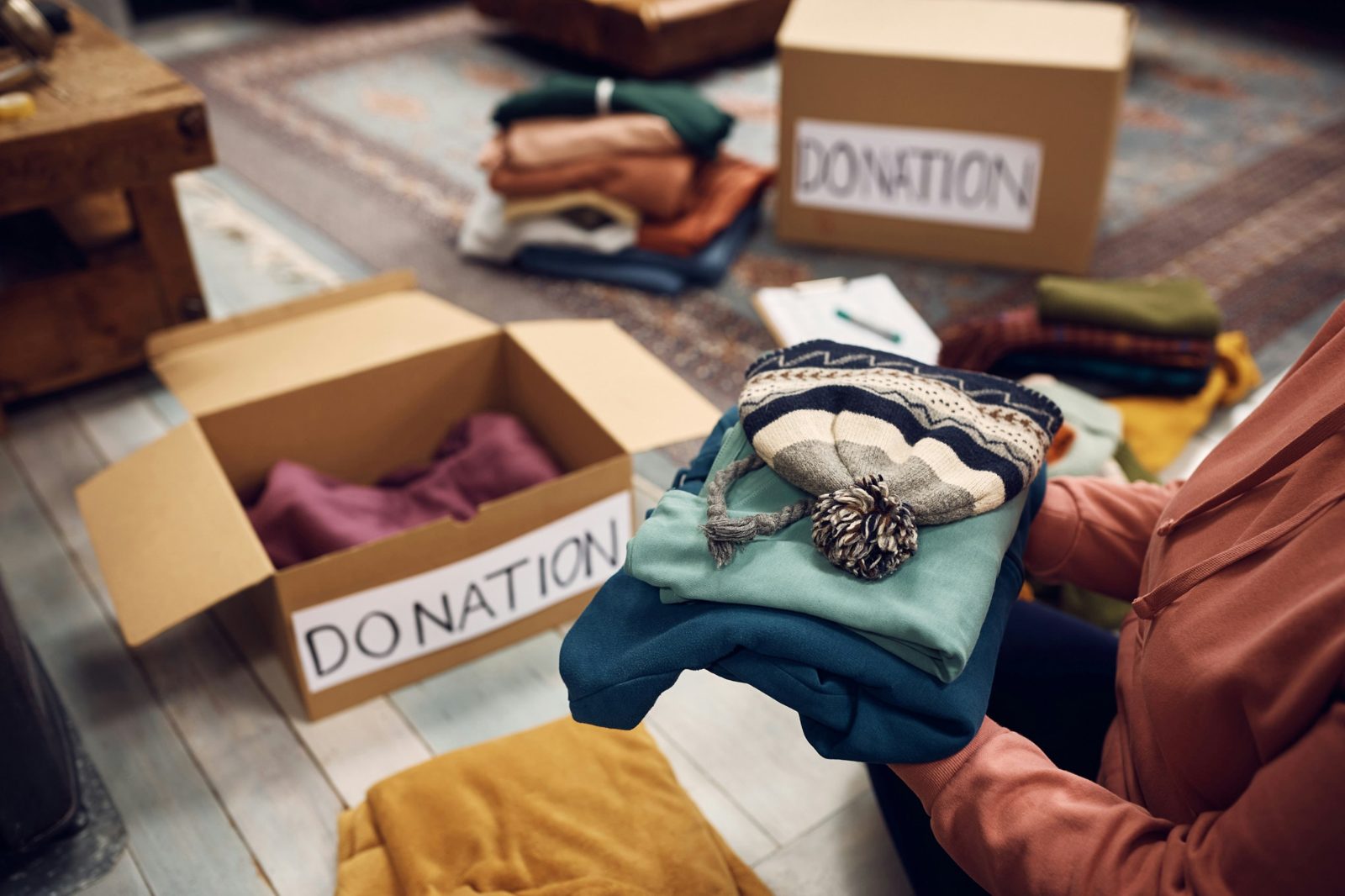 Hands holding folded sweaters and a knit toque beside cardboard boxes marked Donation
