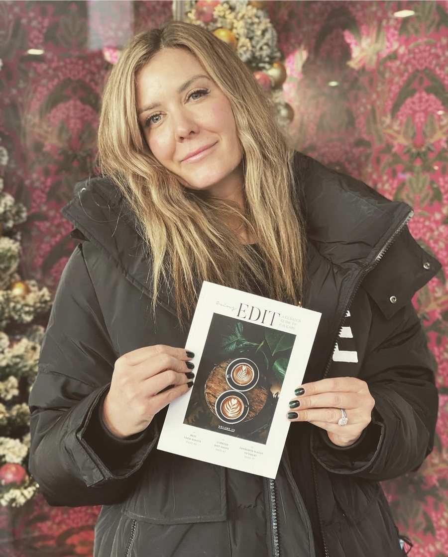 Erin in a winter coat holding Volume III in front of pink floral wallpaper